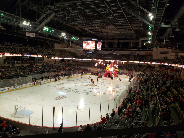 Ein Hockey-Spiel in einer großen Arena mit Zuschauern auf Sitzen und Stehplätzen, umgeben von einem Zaun und Bannern, unter einem Dach mit Metallstangen, Lampen und zwei Bildschirmen, identifiziert als Columbus Blue Jackets Arena.