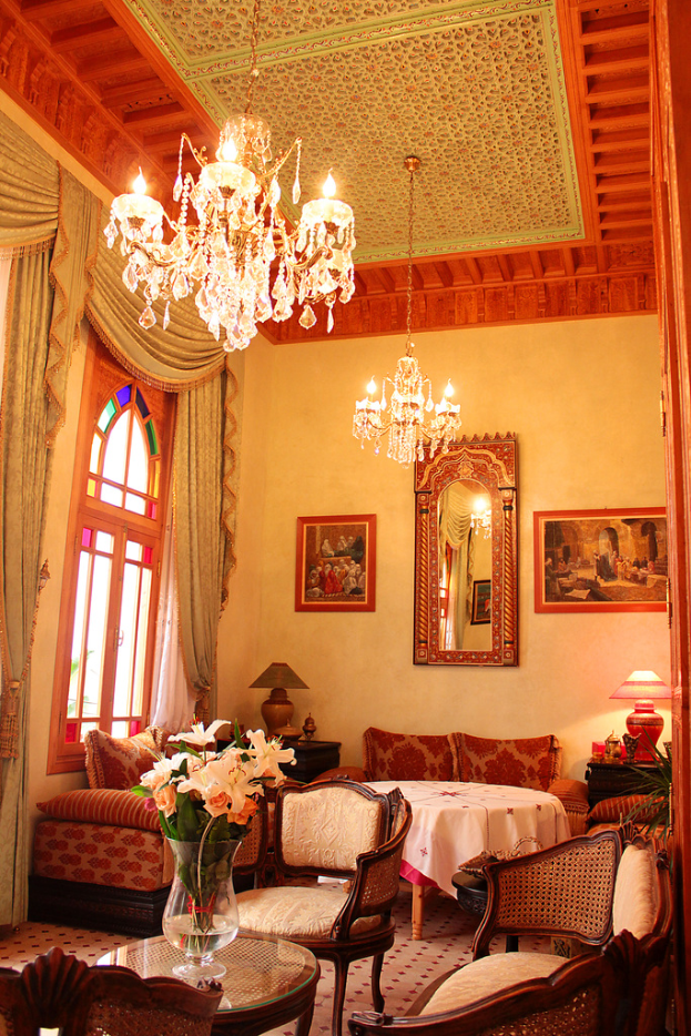 Ein Wohnzimmer mit einem Tisch und Blumentopf im Vordergrund, umgeben von Stühlen, Sofas und einem im Hintergrund mit einer weißen Decke bedeckten Tisch, gerahmte Bilder an der Wand, von der Decke hängende Lampen und Vorhänge mit einem Fenster links.