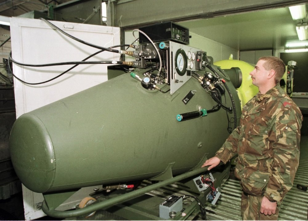 Ein Mann in militärischer Uniform steht neben einer großen grünen Bombe in einem unordentlichen industriellen Setting mit Maschinen, Kabeln und beleuchteten Wandlampen, mit Text unten.