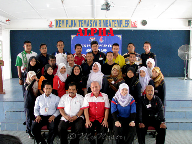 Eine Menschenmenge, die lachend vor einer Bühne posiert, mit einer Wandtafel dahinter, einem Tischventilator rechts, einem Podium links und einem Projektor oben im Bild.