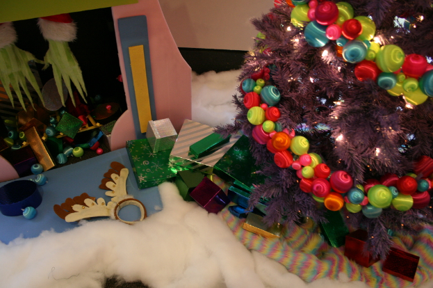 Drinnen steht rechts ein Weihnachtsbaum mit Lichterketten und Schmuck, in der Mitte Geschenkboxen und Deko, links oben eine Hütte mit Deko und unten Baumwolle und Wolle.