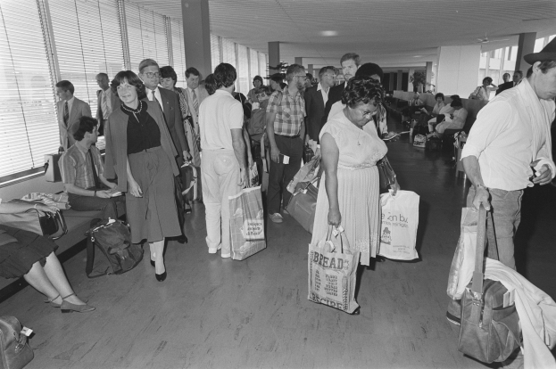 Schwarzes und weißes Bild von Reisenden in einem Flughafen, einige sitzen auf Bänken, andere stehen mit Gepäck, mit einem Mann und einer Frau, die Taschen halten, in der Mitte und Fensterläden links.