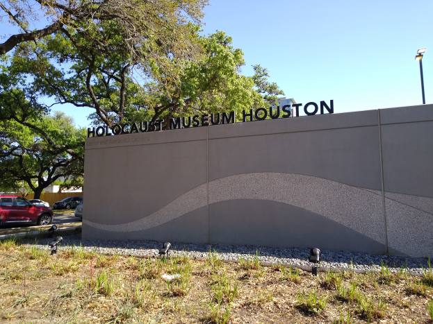 Außenansicht des Houston Holocaust Museums mit eingravierter Textwand, Gedenksteine, Landschaftsgestaltung, vorbeifahrende Fahrzeuge und einem bewölkten Himmel.