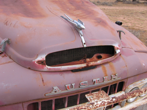 Ein stark verrosteter, alter Lkw auf einer grünen Wiese mit Bäumen und einem klaren blauen Himmel im Hintergrund.