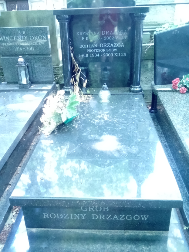 Ein Friedhof mit zwei benachbarten Grabsteinen umgeben von Blumensträußen und anderen Gegenständen, mit einem Gebäude im Hintergrund.