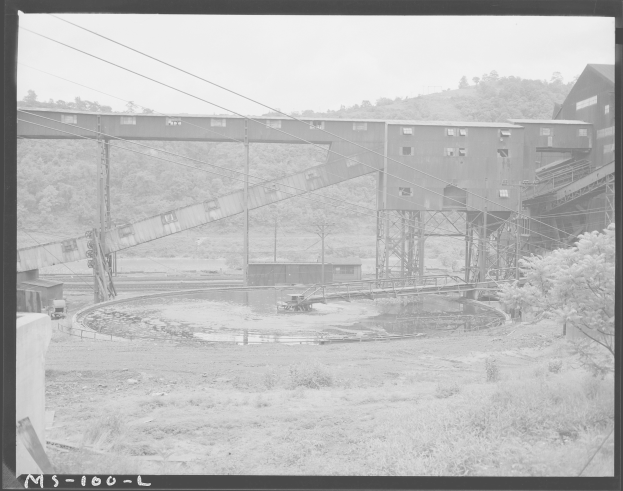 Ein Schwarz-Weiß-Foto eines Kohlebergwerks mit einer Brücke, Polen, Drähten, Gebäuden, Bäumen, Pflanzen, Gras und Wasser im Vordergrund und einem Himmel im Hintergrund sowie Text unten.