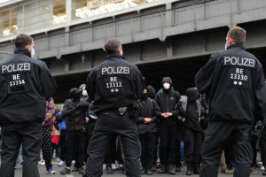 Eine Gruppe uniformierter Polizisten steht vor einer Menge von Menschen in schwarzen Uniformen und Masken, mit einer Brücke und einem Gebäude im Hintergrund, was eine städtische Umgebung vermuten lässt.