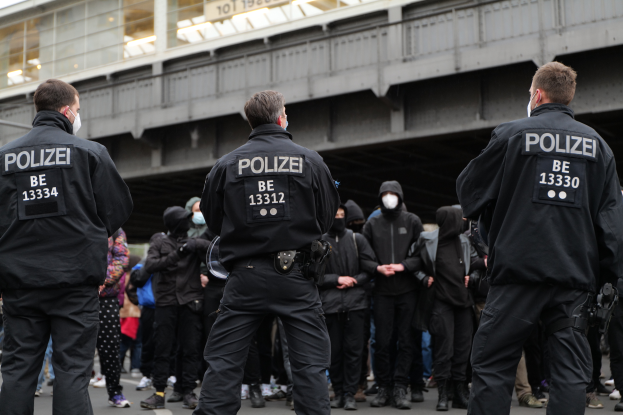 Eine Gruppe uniformierter Polizisten steht vor einer Menge von Menschen in schwarzen Uniformen und Masken, mit einer Brücke und einem Gebäude im Hintergrund, was eine städtische Umgebung vermuten lässt.