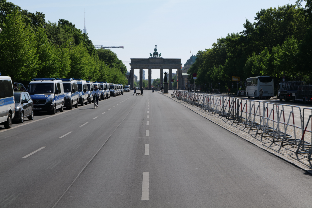 Eine Reihe von Polizeiwagen, die auf einer Straße vor dem Brandenburgertor in Berlin, Deutschland, geparkt sind, mit Menschen auf Fahrrädern und Stehenden, Barrieren, Bäumen und einem Bogen mit Statuen im Hintergrund.