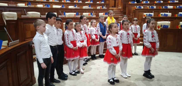 Eine Gruppe von Kindern in weißen und roten Kleidern steht zusammen in einem Raum, mit einem Podium und einem Mikrofon auf der linken Seite und Stühlen und Tischen mit verschiedenen Gegenständen im Hintergrund, die Flaggen tragen, die ihre Teilnahme an einer Nationalversammlung anzeigen.