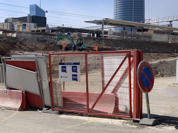Baustelle mit rotem Tor und Schild, Metallzaun, Gebäuden, Kränen, Polen, Drähten, wolkenlosem Himmel und Arbeitern mit Schildern im Hintergrund.