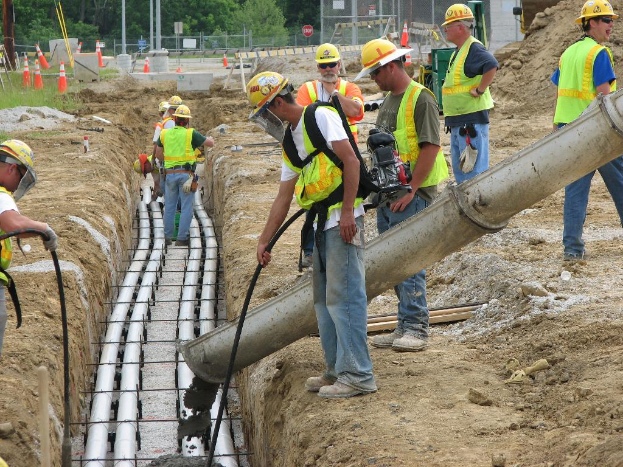 Eine Baustelle mit Arbeitern in Jacken und Helmen, Absperrbaken, Zaun, Bäumen, Pfählen, Erde und Rohren im Hintergrund.