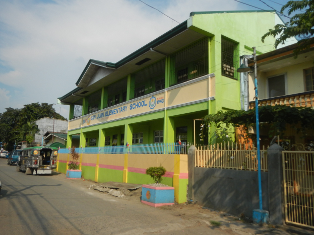 Schulgebäude mit grüner und gelber Fassade, Fahrzeuge auf der Straße, Topfpflanzen, ein Zaun, ein Tor, ein Straßenpfahl, Bäume, Drähte und ein bewölkter Himmel.