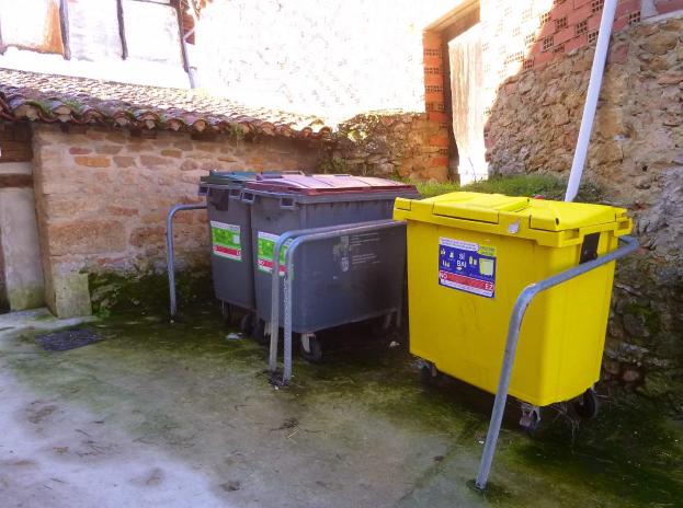Zwei gelbe und graue Mülltonnen mit Rollen nebeneinander vor einem Gebäude mit einer Wand und einem Fenster im Hintergrund.