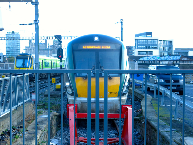 Zwei Züge auf den Schienen mit einem Zaun im Vordergrund, einige Pfosten auf dem Land, einer mit einer Signalleuchte, ein Auto auf der Straße rechts, Gebäude im Hintergrund und der Himmel darüber.