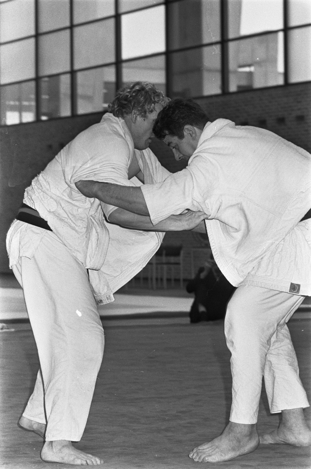 Zwei Männer in weißen Judo-Anzügen kämpfen heftig auf einem Boden mit einer Wand und Glasfenstern im Hintergrund, dargestellt in Schwarz-Weiß.