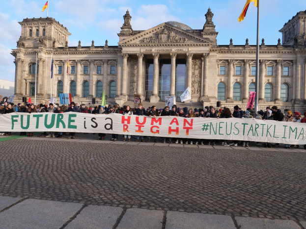 Eine Gruppe von Menschen hält ein Banner mit der Aufschrift "Zukunft ist ein Mensch" vor dem Reichstaggebäude in Berlin, Deutschland, mit sichtbaren architektonischen Details und Fahnen im Hintergrund unter einem bewölkten Himmel.