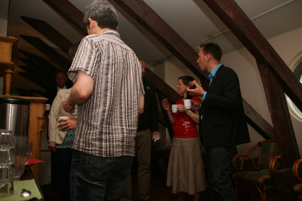 Mehrere Personen mit Tassen in der Hand, mit einer Tasse und einem Tisch auf der linken Seite, einem Dach oben und Stühlen auf der rechten Seite.