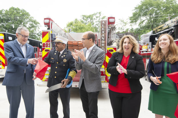 Eine Gruppe von Menschen bei einer Eröffnungszeremonie für die Alexandria Fire Department, die vor einem Feuerwehrauto klatschen und lächeln, während zwei eine Schere und ein rotes Band halten.