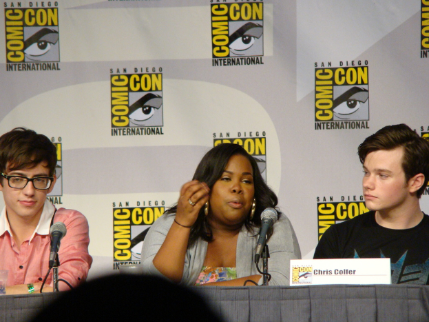 Eine Gruppe von Menschen, die an einem Tisch mit Mikrofonen, einer Namensschilder und einer Werbetafel im Hintergrund sitzen, wobei ein Mann eine Brille trägt.