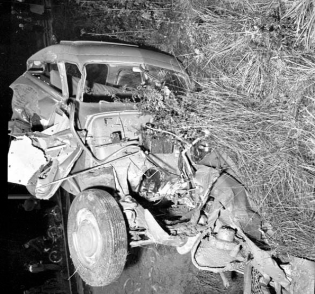 Ein Schwarz-Weiß-Foto eines schwer beschädigten Autos am Straßenrand mit einer zerquetschten Front, zersplitterten Fenstern, einem Baum im Hintergrund und verstreuten Gegenständen.