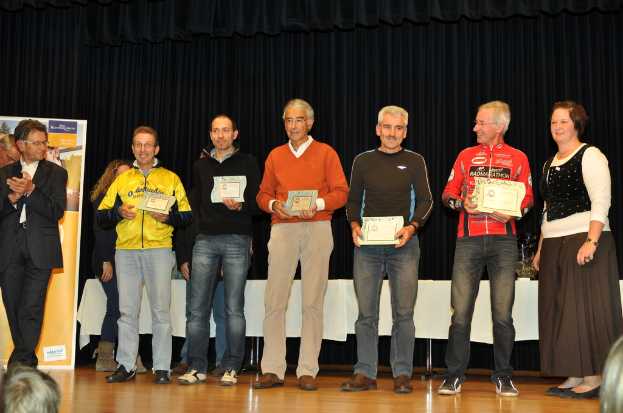 Eine Gruppe von Menschen steht auf einer Bühne und hält Zertifikate in der Hand, mit einem Banner im Hintergrund und einem Tisch daneben, alles vor einem schwarzen Vorhang.