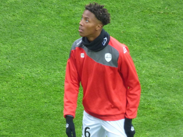 Ein junger Mann in roter Jacke und weißen Shorts steht auf einem Fu�ballfeld und schaut rechts mit entschlossener Miene, mit Baseballhandschuhen ausgestattet.