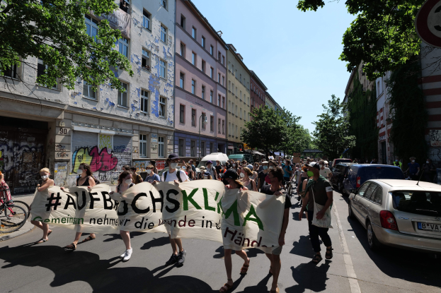 Eine Gruppe von Menschen marschiert auf einer Straße in Berlin, hält ein Banner in der Hand, mit parkenden Autos auf der rechten Seite und einem Fahrradfahrer auf der linken Seite, vor einem Hintergrund von Gebäuden, Bäumen und einer Schautafel.