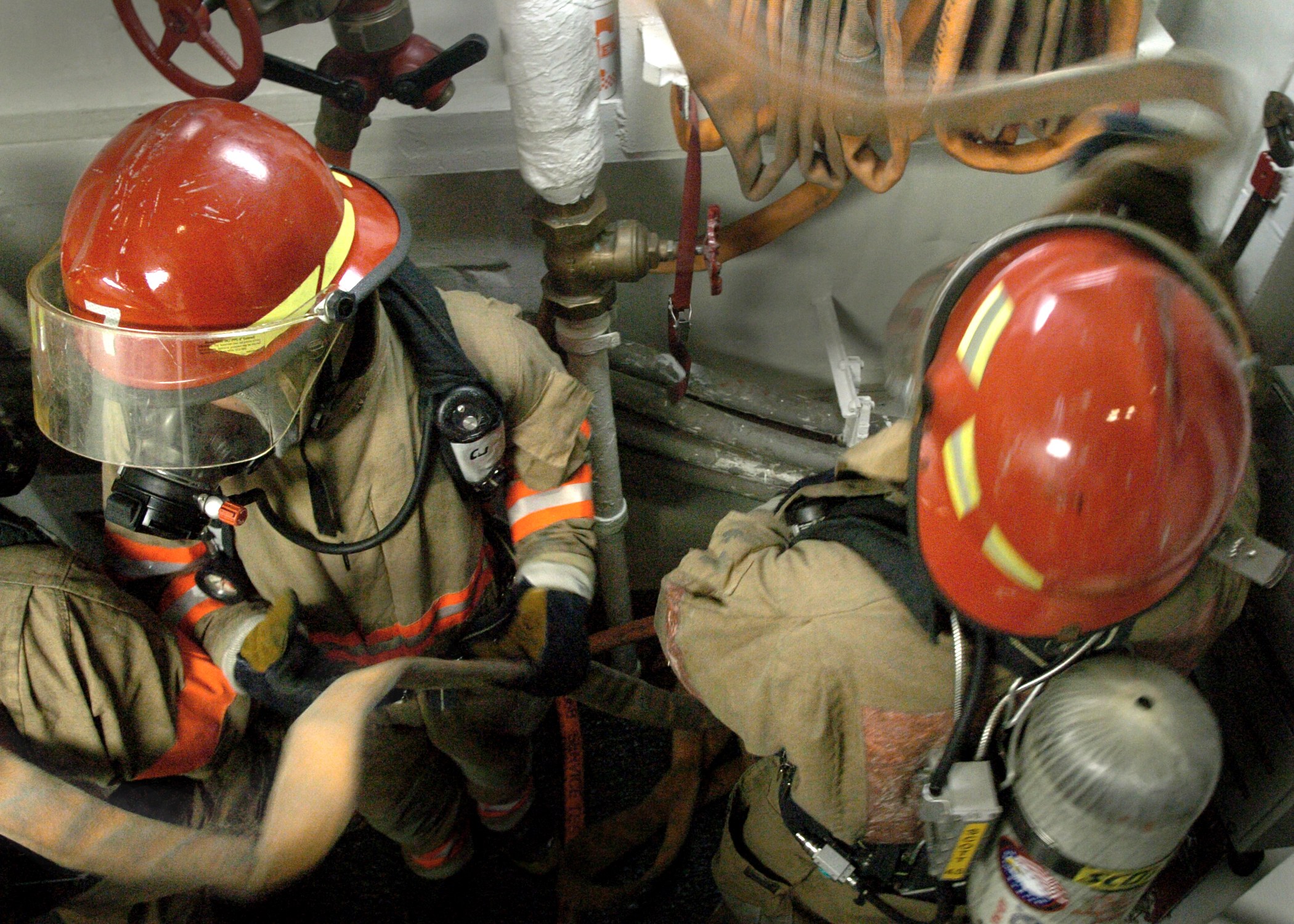 Feuerwehrleute in Schutzausrüstung arbeiten an einem Feuerhydranten mit Rohren und Ventilen im Hintergrund.