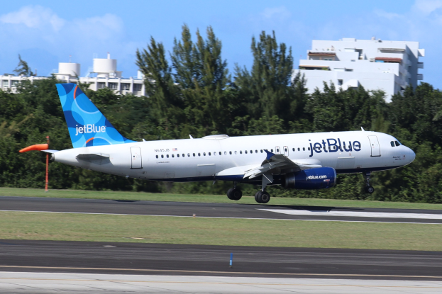 JetBlue Airbus A320-200 beim Start von Miami International Airport mit grünem Gras, hohen Bäumen, Gebäuden im Hintergrund und einem hellblauen Himmel.