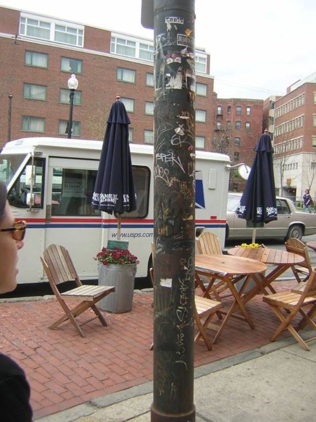 Ein Tisch und Stühle sind auf einem Gehweg aufgestellt, mit Autos und Lieferwagen auf der benachbarten Straße geparkt und Gebäude im Hintergrund sichtbar.