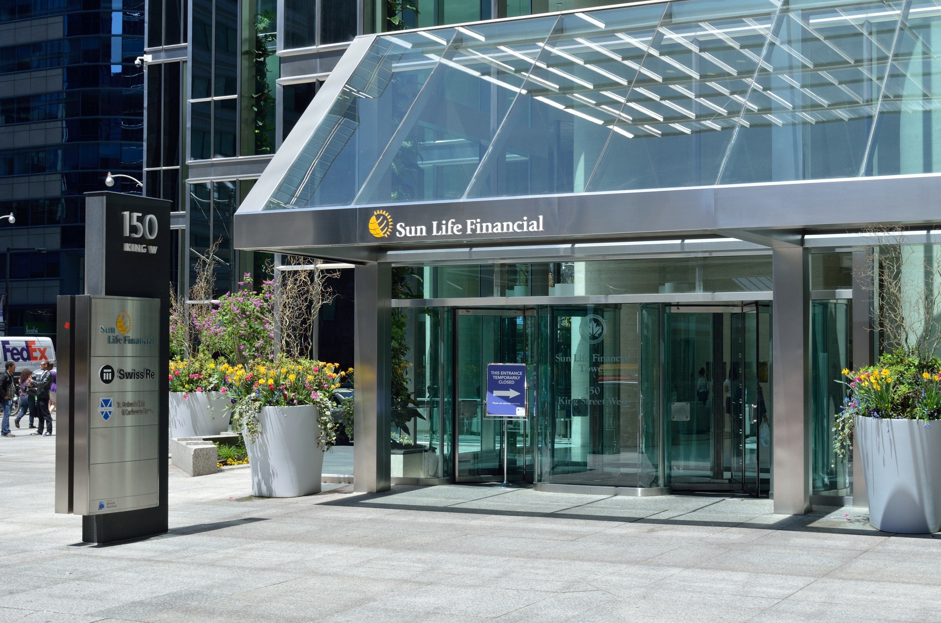 Moderner gläserner Sun Life Financial-Gebäude in Toronto mit einem Schild, Topfpflanzen mit Blumen, Passanten, einem Straßenschild, einem Schild, Fahrzeugen auf der Straße und einer bewölkten Himmel.