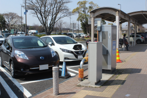 Ladestation für Elektroauto in Japan mit Autos, Verkehrskegeln, einer Person auf dem Gehweg, einem Schuppen, Mästen, Lichtern, Schildern, Bäumen, Pflanzen und einem Himmel im Hintergrund.