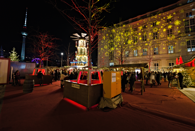 Ein geschäftiger Weihnachtsmarkt in Berlin, Deutschland, mit Menschen um beleuchtete Stände, Bäume, Gebäude, Laternenpfähle und einen Turm im Hintergrund unter einem dunklen Himmel.