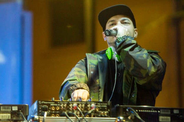 Ein Mann mit Mütze und Kopfhörern singt in ein Mikrofon, während er einen DJ-Set spielt, umgeben von elektronischen Geräten mit leicht unscharfem Hintergrund.