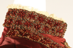 Nahaufnahme eines roten und goldenen Kleides mit detaillierter Spitzenverzierung auf einem Mannequin vor einem weißen Hintergrund.