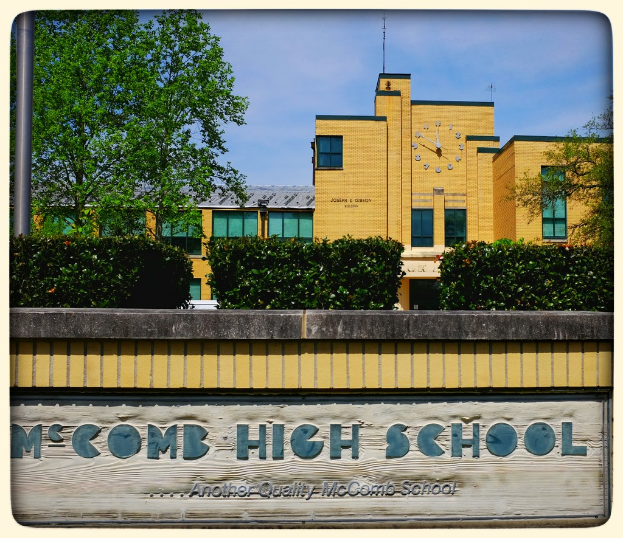Ein Plakat mit der Aufschrift "MCCOMB HIGH SCHOOL" auf einer Wand mit Pflanzen, Bäumen, einem Pfahl, einem Gebäude und einem bewölkten Himmel im Hintergrund.