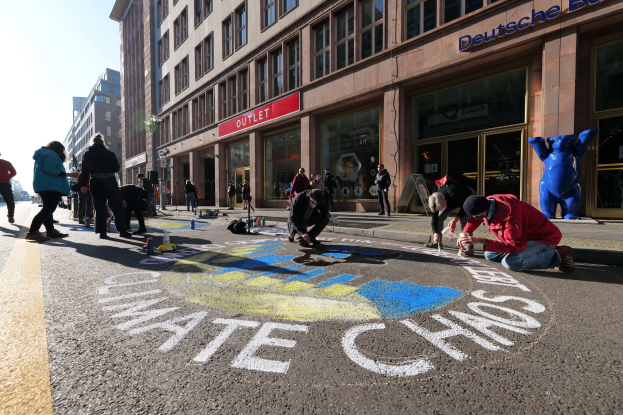 Eine Gruppe von Menschen sitzt vor einem Gebäude mit Fenstern und Namensschildern auf dem Boden, umgeben von Flaschen und anderen Gegenständen, während sie an einer Klimawandel-Demonstration in Berlin teilnehmen, mit Bäumen und einem klaren blauen Himmel im Hintergrund.