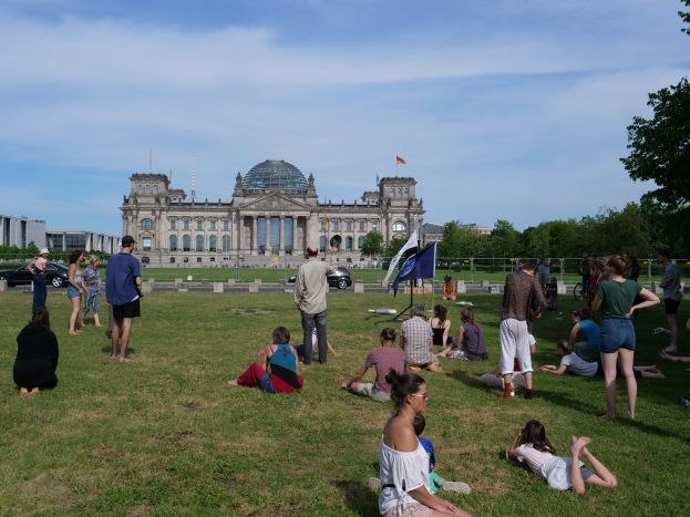 Eine Gruppe von Menschen sitzt auf einem grünen Feld vor dem Reichstaggebäude in Berlin, Deutschland, umgeben von Bäumen, Fahrzeugen und Fahnen auf Stangen unter einem bewölkten Himmel.