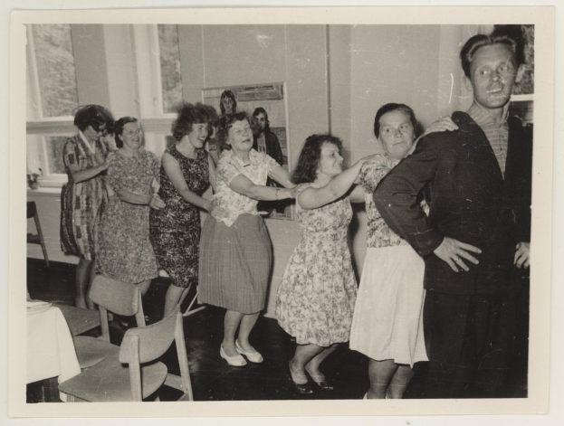Gruppe von Menschen, die in einem rückw├Ąrtsgewandten Raum mit Stühlen, einem mit einer wei├čen Decke bedeckten Tisch, einem Foto auf der Wand und Fenstern im Hintergrund tanzen.