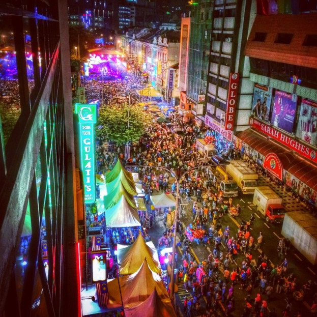Ein lebhaftes Karnevalsszenario mit einer großen Menschenmenge auf der Straße, umgeben von Zelten, Geschäften, Gebäuden und Bannern, mit zahlreichen Lichtern im Hintergrund.