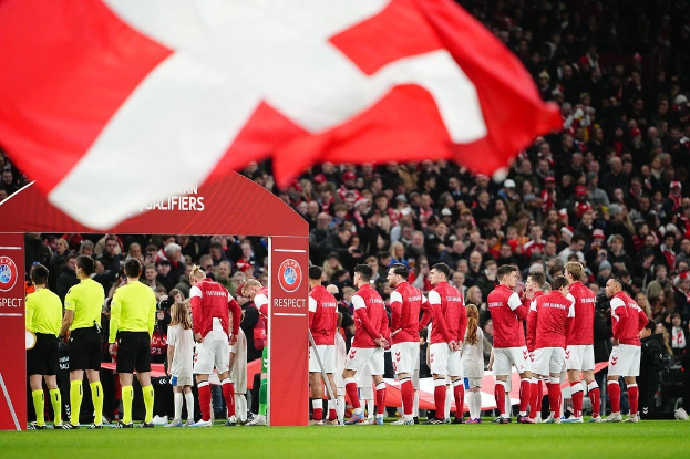 Eine Gruppe von Menschen auf einem Fussballfeld mit einer roten und weißen Flagge im Vordergrund, einem Bogen mit der Aufschrift "Bayern München vs Bayern München Wetten & Vorschau" im Hintergrund und einer großen Menge im Stadion.