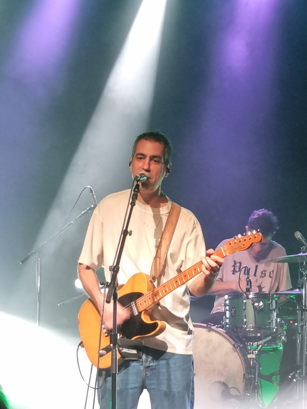 Ein Mann spielt Gitarre auf der Bühne, beleuchtet von Scheinwerfern von oben, während er in ein Mikrofon singt, mit einem anderen Mann, der im Hintergrund Schlagzeug spielt.