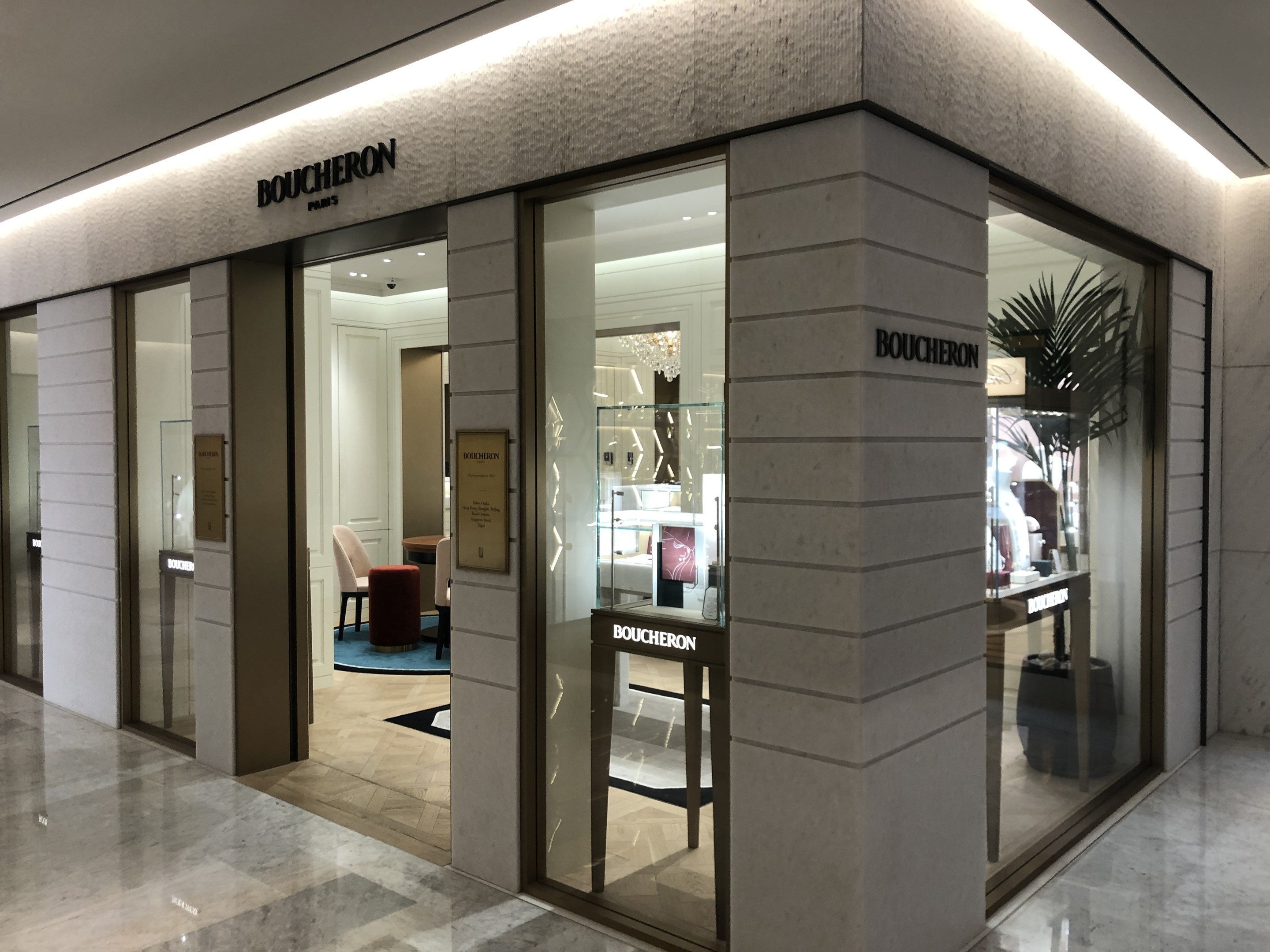 Eintritt zu einem Boutique in einem Einkaufszentrum mit Glaswänden, Türen, Text an den Wänden, Deckenleuchten, Stühlen, Tischen, einer Pflanze und sichtbarem Boden.