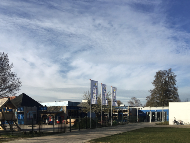 Großes Gebäude mit einem Metallzaun, Rasenfläche, Weg, Bäumen, Bannern, einem Fahrrad, nahen Häusern und einem bewölkten Himmel, identifiziert als der neue Spielplatz an der Schule.