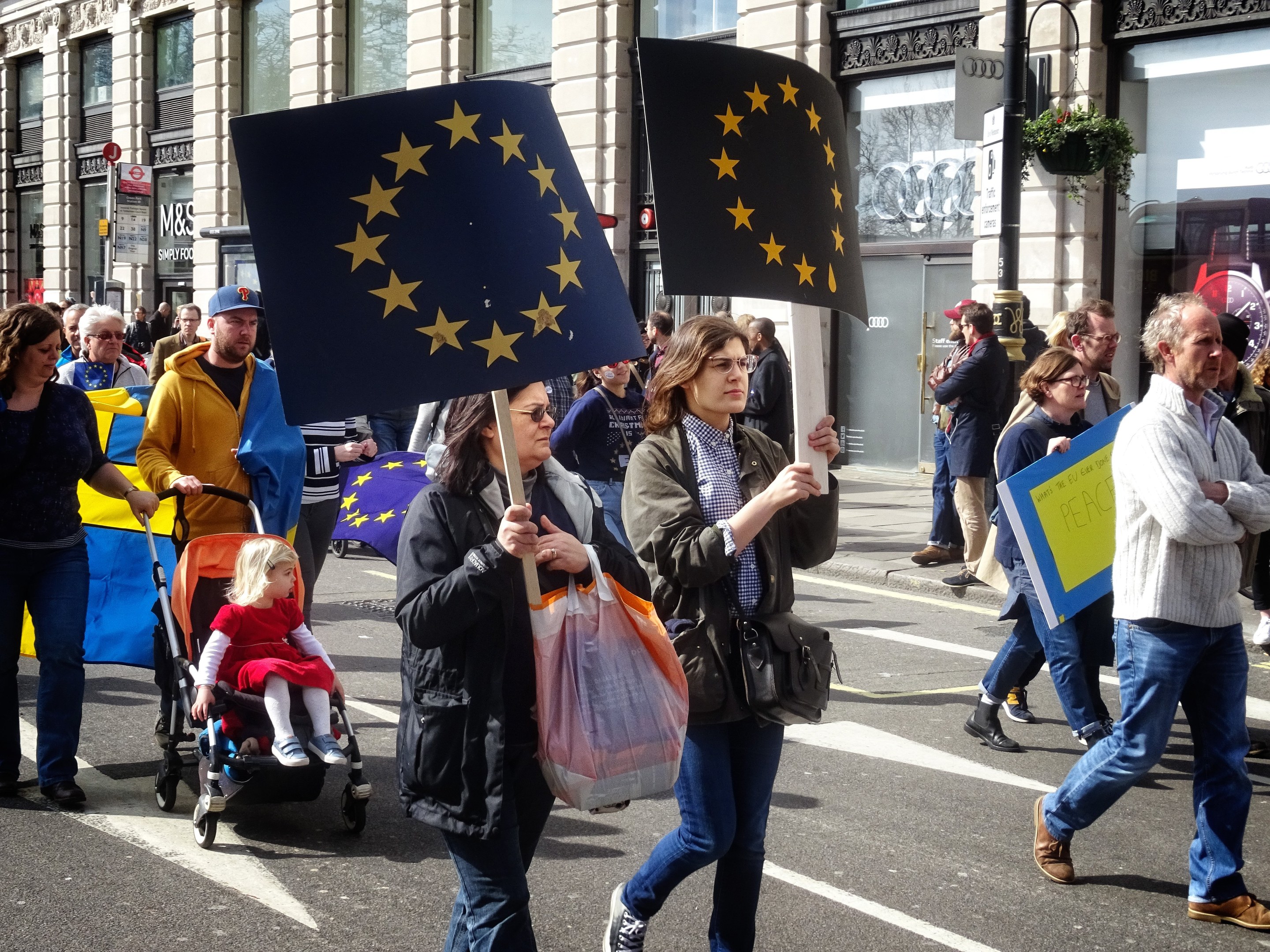 Eine Gruppe von Menschen, die eine Straße entlanggehen, einige halten Fahnen und Schilder, mit einem Baby im Kinderwagen und Gebäuden, Schildern und Topfpflanzen im Hintergrund.