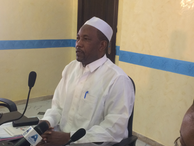 Ein Mann in einem weißen Kleid und Hut sitzt an einem Tisch mit Mikrofonen, spricht, während Papiere und Gegenstände auf dem Tisch zu sehen sind, mit einem anderen Menschen mit Brille rechts von ihm, in einem Raum mit einer Wand und einer Tür im Hintergrund.