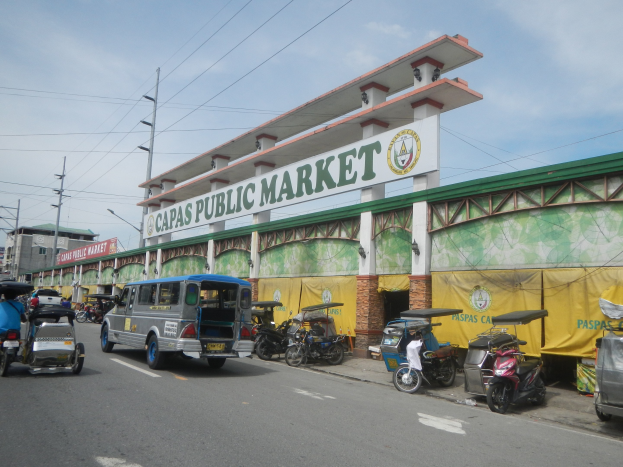 Eine belebte Stadtstraße mit Autos, Motorrädern und Rikschas, die an einem Gebäude mit der Aufschrift "Capas Public Market" vorbeifahren, vor einem wolkenverhangenen Himmel mit Strommasten und Gebäuden im Hintergrund.
