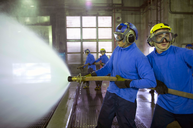 Eine Gruppe von Männern in blauen Hemden und gelben Helmen arbeitet an einer Maschine in einer Fabrikumgebung, wobei einer der Männer ein Rohr hölt und Wasser auf den Boden spröht.