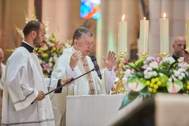 Der Bischof der Kirche des Heiligen Grabes steht an einem Rednerpult mit einem Mikrofon und spricht zu einer Gruppe von Menschen. Rechts von ihm befindet sich ein Tisch mit Kerzen und Blumen, im Hintergrund gibt es Blumensträuße und ein Buntglasfenster.